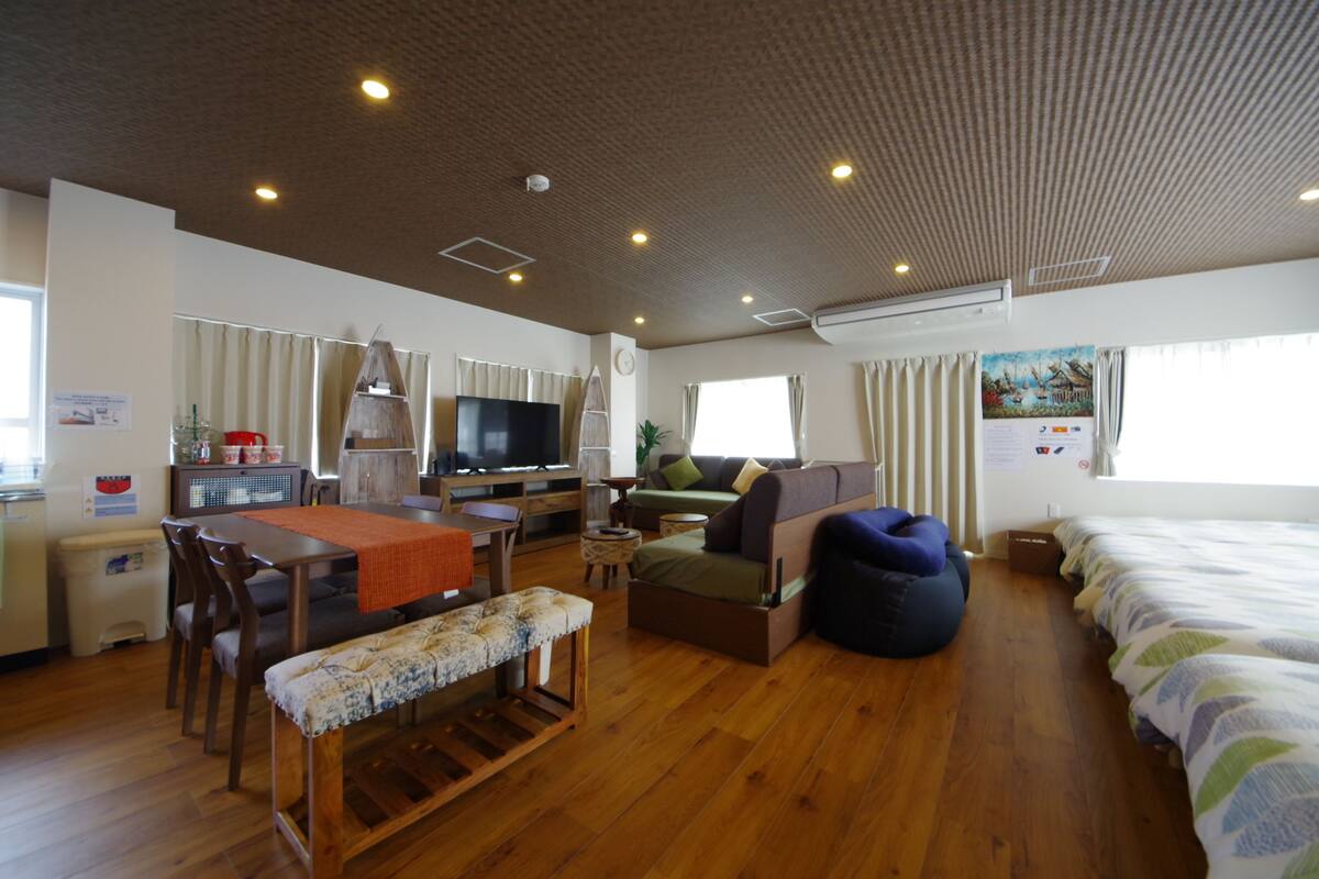 Quarto bem iluminado com piso de madeira, mesa de jantar com cadeiras, sofá, cama e televisão.