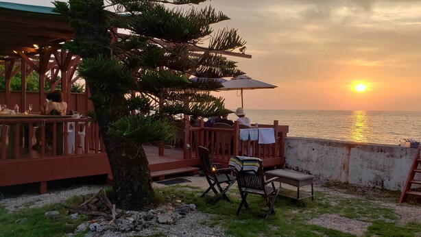 A coastal restaurant with outdoor seating overlooking the sea, featuring a wooden deck and a view of the sunset. - Beach Plus (Onna)