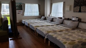 A hotel room with four single beds, a wooden floor, a window with curtains, a wall-mounted artwork, and a small table with a lamp. - Beach Plus (Onna)