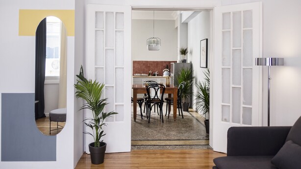 A modern living room with a black sofa, a dining area with a wooden table and chairs, and a kitchen in the background. - Live Life Ermou (Athens)
