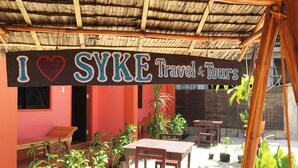 A sign reading "I ♥ SYKE Travel & Tours" above a wooden table and chairs in an outdoor setting with potted plants.