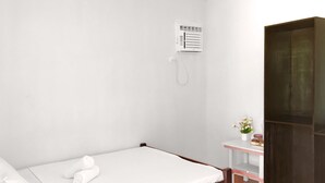 A single bed with white bedding, a small bedside table with a vase of flowers, and a wall-mounted air conditioning unit.