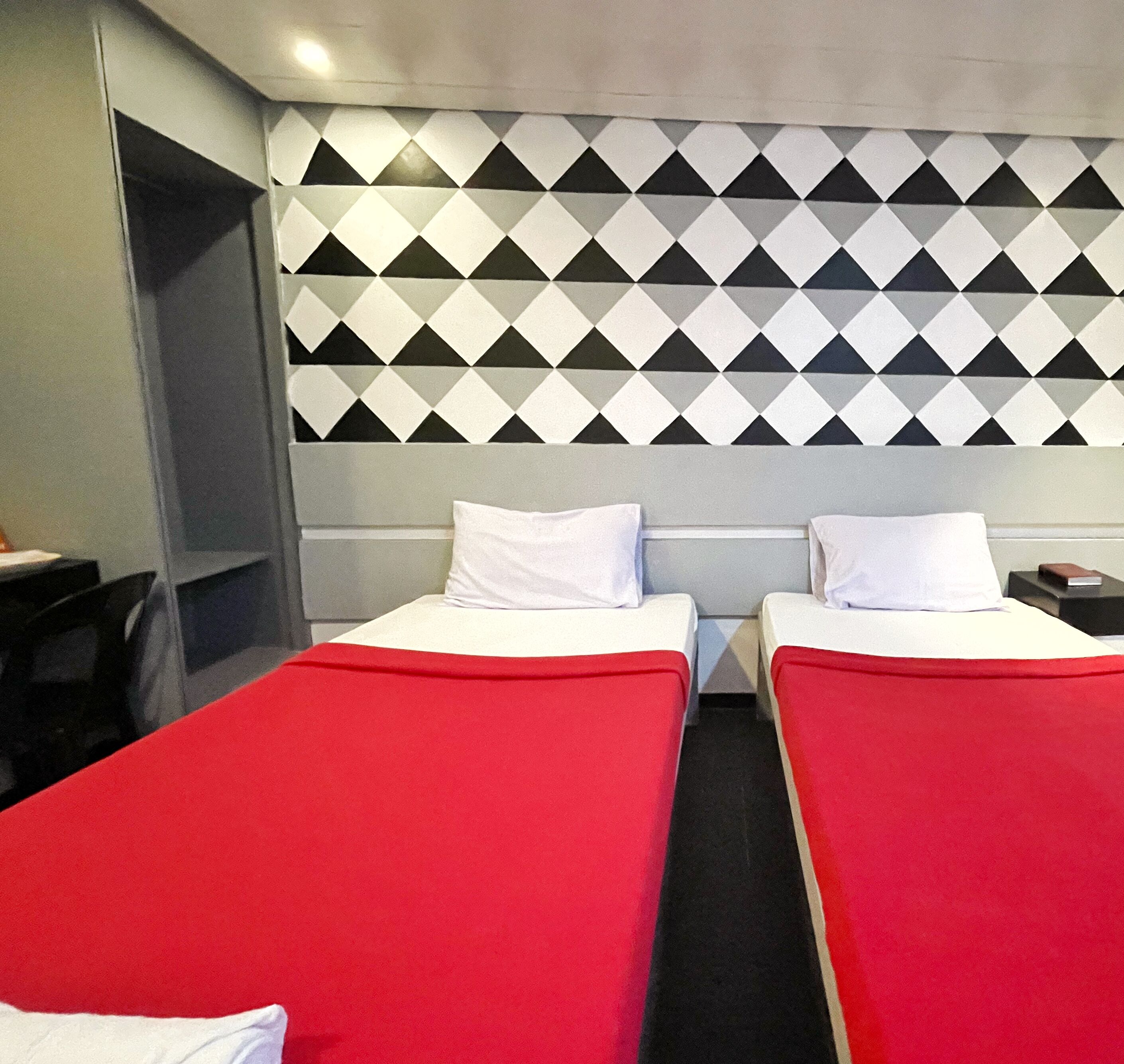 Quarto de hotel com duas camas de solteiro, cada uma com colcha vermelha e almofadas brancas. A cabeceira apresenta um padrão geométrico em preto e branco.