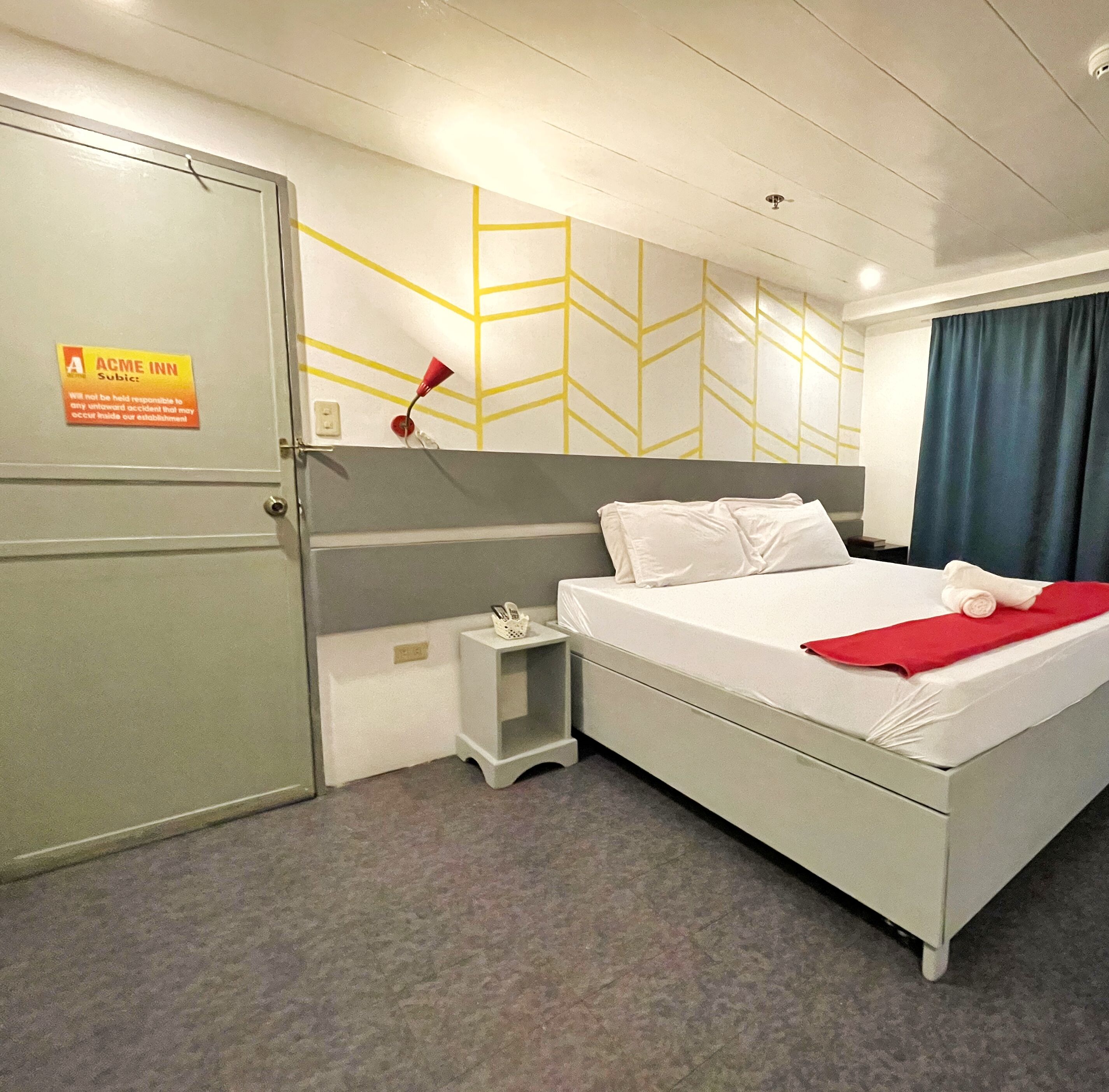 Habitación de hotel con cama, mesita de noche, teléfono, toalla roja y un cartel en la puerta.