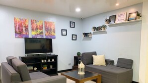 A living room with a flat-screen TV, grey sofas, a coffee table, and decorative items including wall art and shelves with plants. - ACME Inn Subic (Olongapo)
