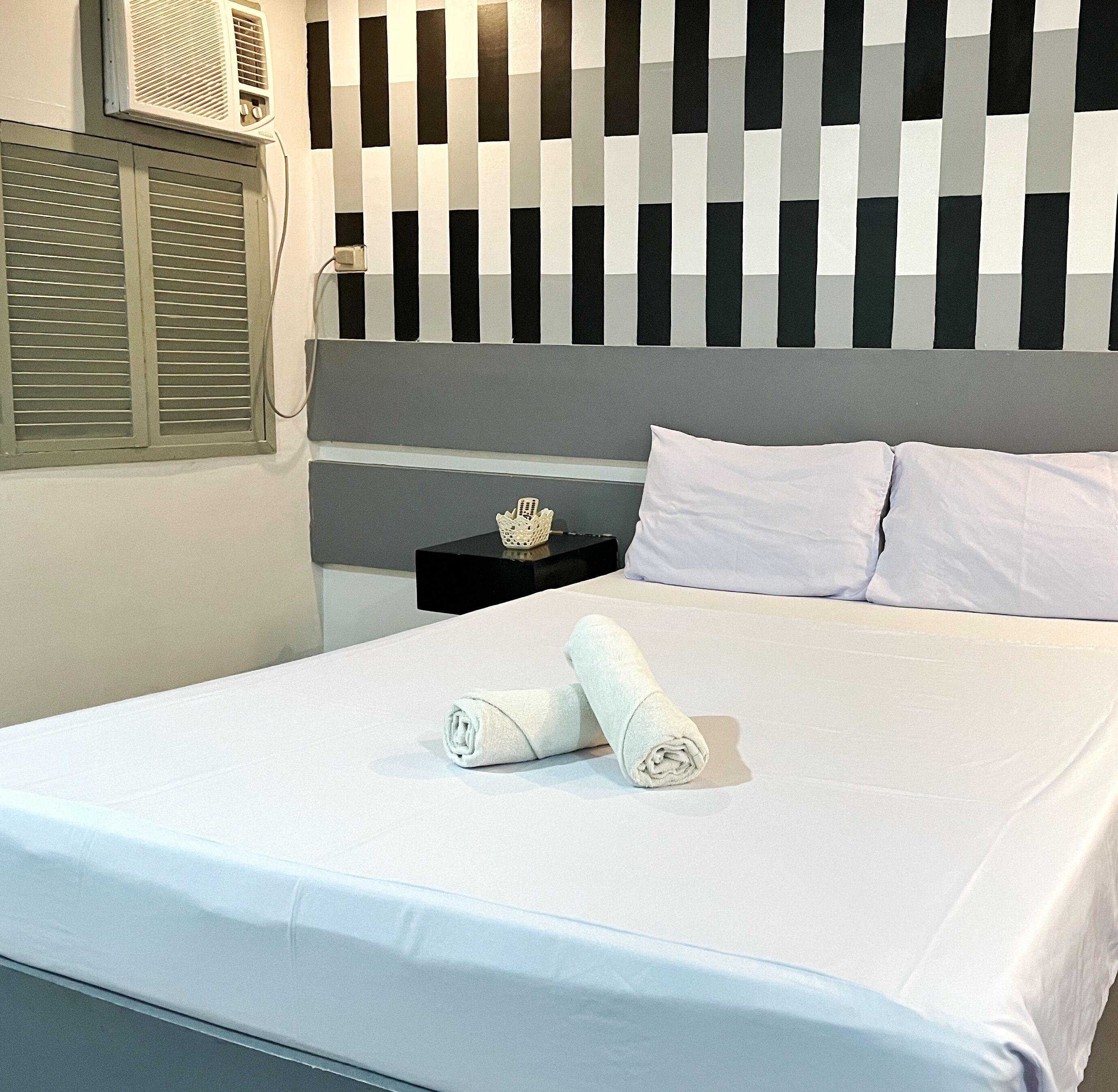 Una cama bien hecha con sábanas blancas y dos toallas enrolladas, una mesita de noche negra con un objeto decorativo, y una pared con estampado a rayas negras y blancas.