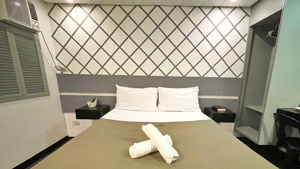 A hotel room with a bed, a headboard with a diamond pattern, a bedside table with a book, and a small table with a chair. - ACME Inn Subic (Olongapo)