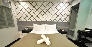 A hotel room with a bed, a headboard with a diamond pattern, a bedside table with a book, and a small table with a chair. - ACME Inn Subic (Olongapo)
