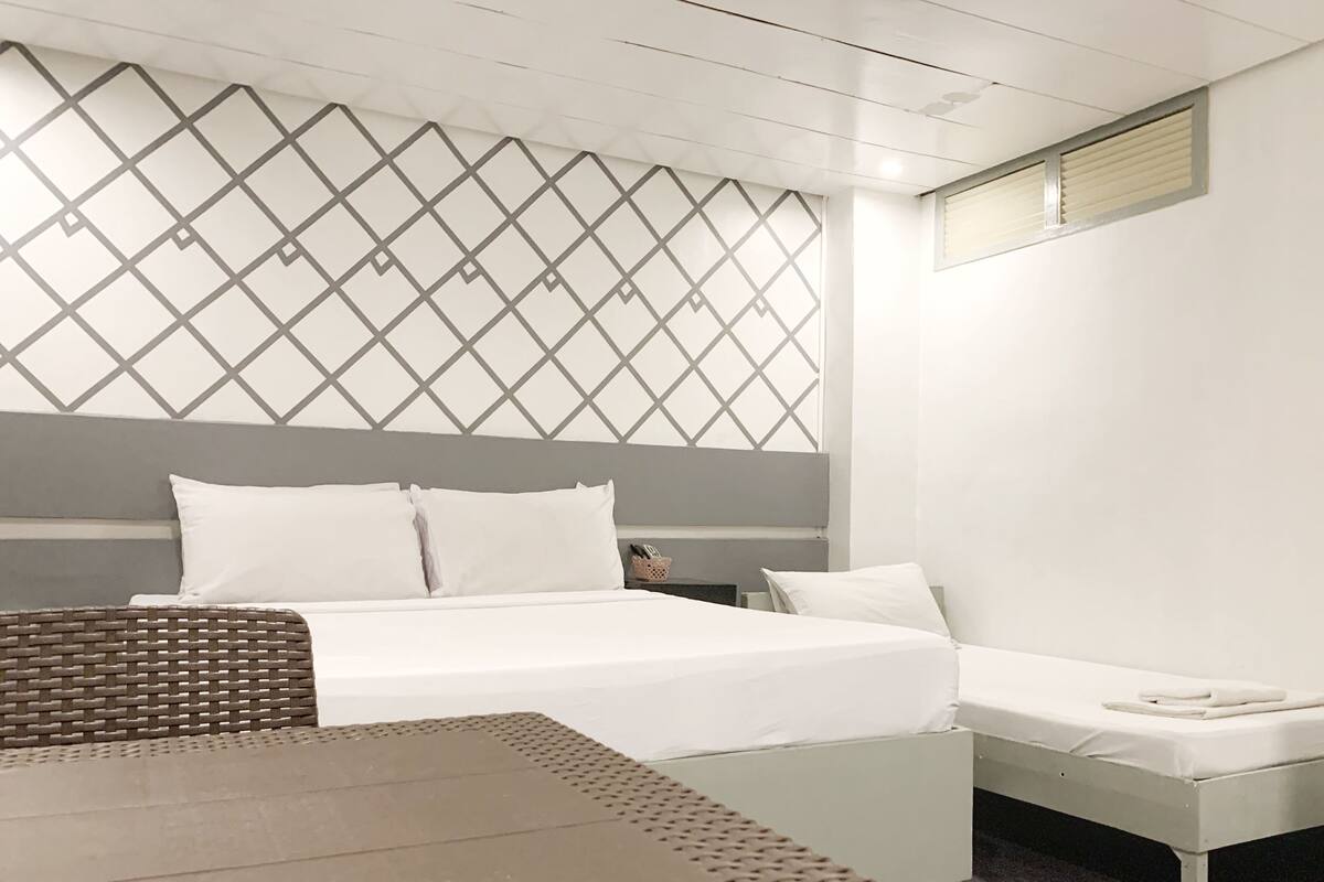Quarto de hotel moderno com uma cama grande, uma mesinha de cabeceira, uma cadeira de vime e um painel decorativo na parede.