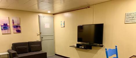 A small, furnished room with a sofa, a television mounted on the wall, a table with a chair, and a door with a sign.