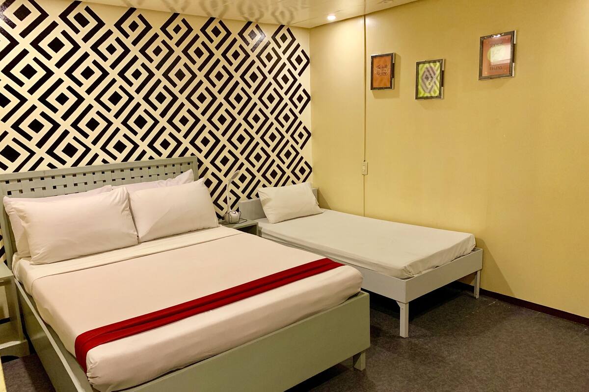 Quarto com uma cama e um sofá-cama pequeno. Há uma parede estampada atrás da cama e uma parede lisa com duas molduras.