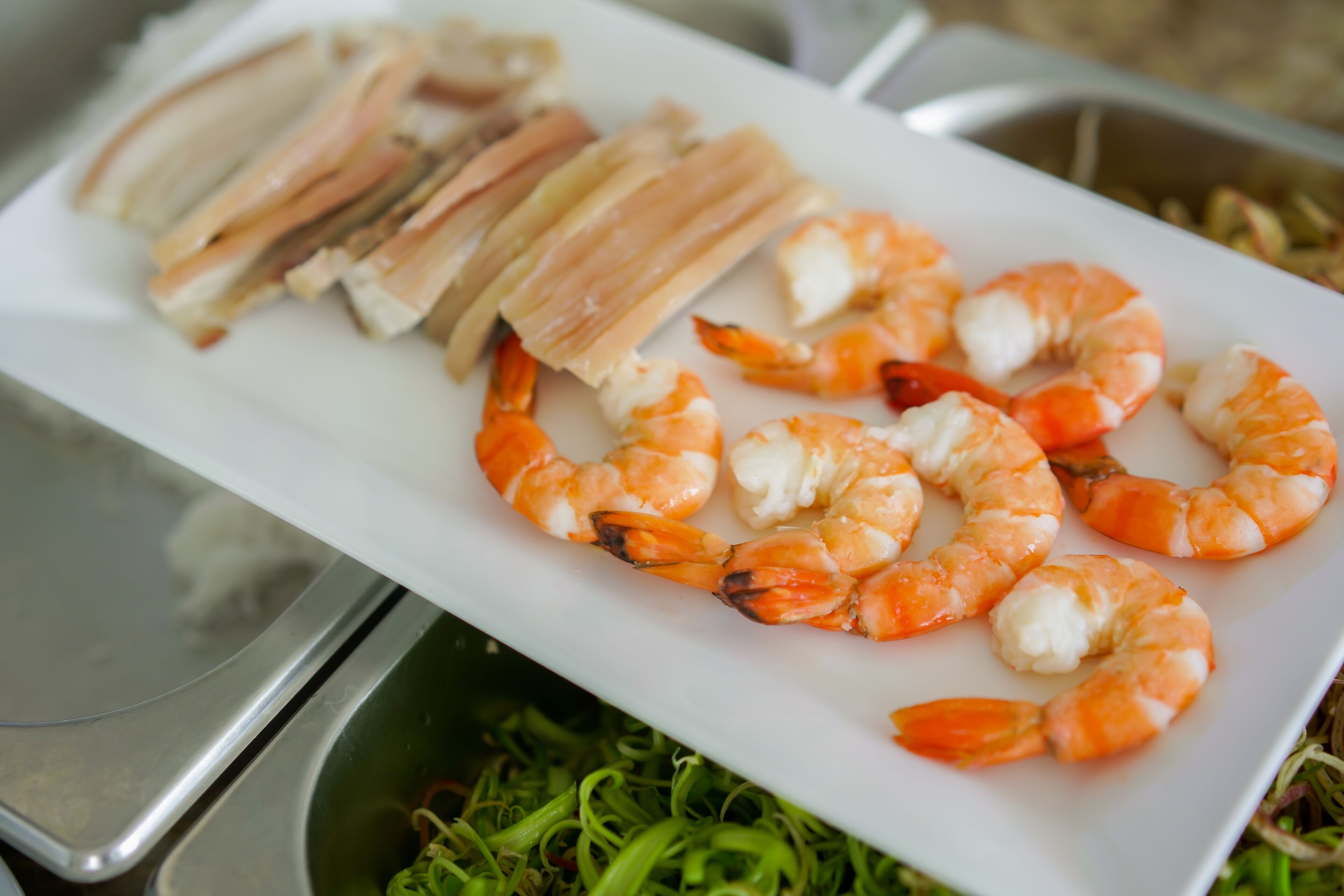A white rectangular plate with cooked shrimp and slices of meat, possibly pork, on a stainless steel table.