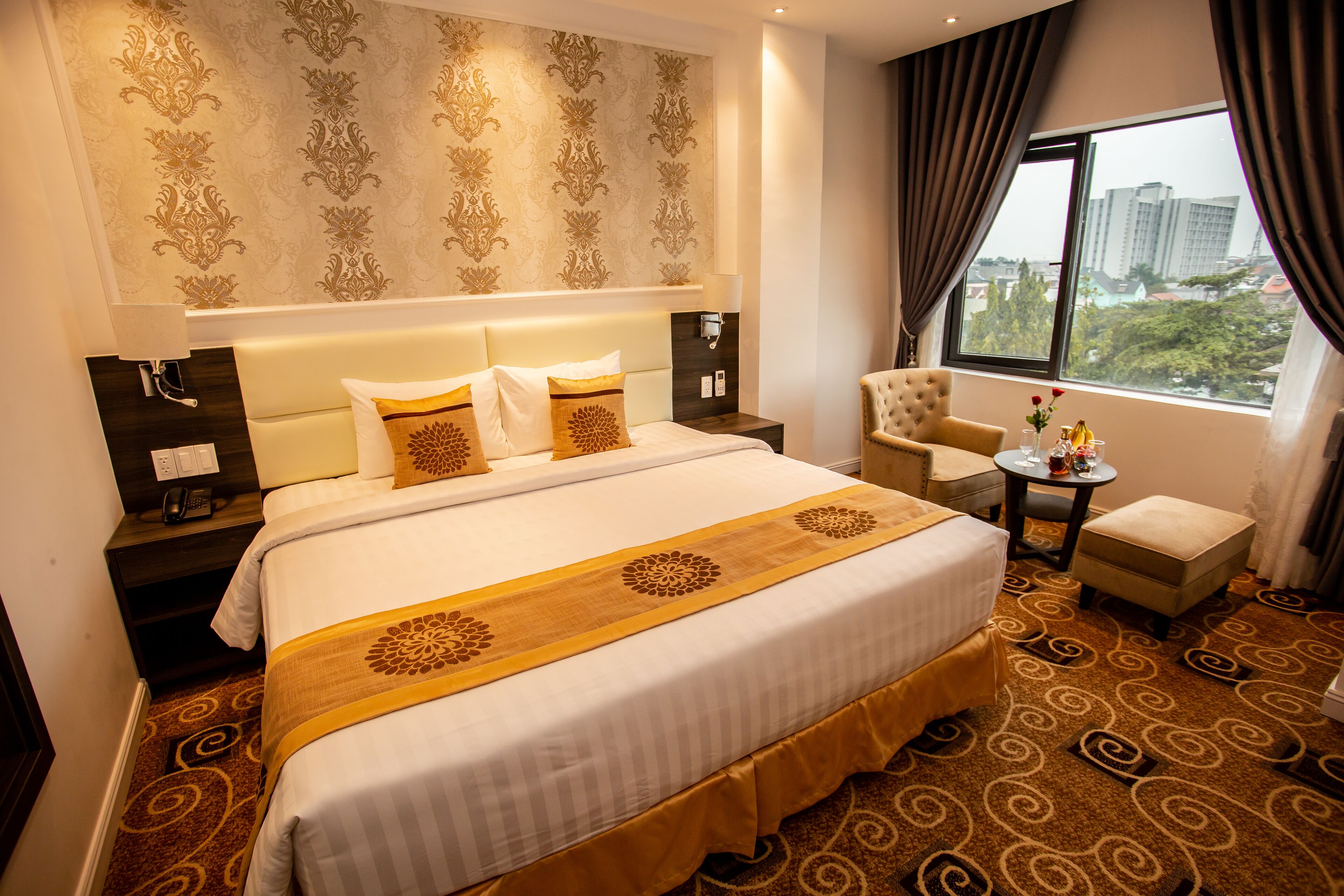 A hotel room with a large bed, a chair, a small table, and a view of the city through the window.