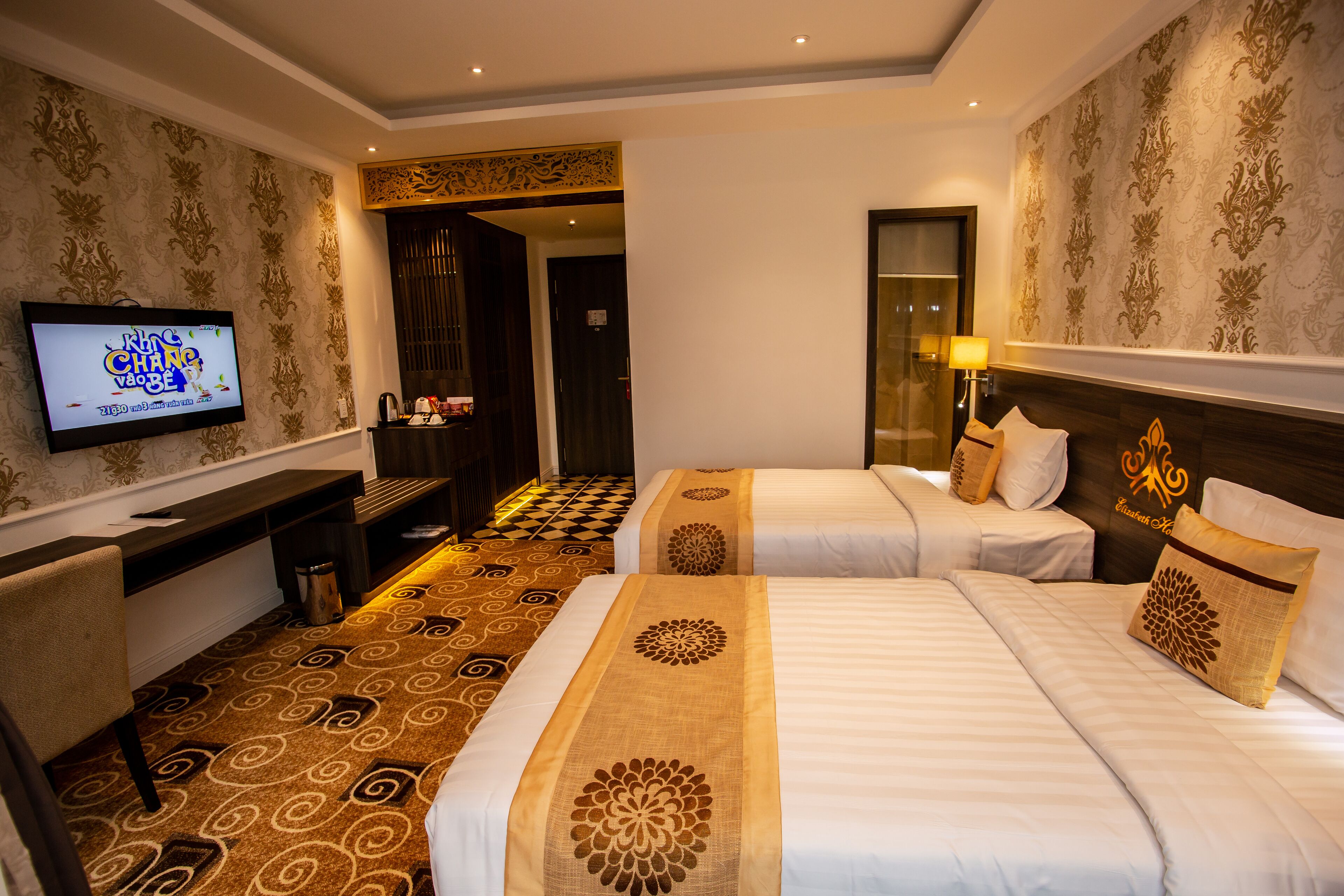 A hotel room with two beds, a TV, a desk, and a patterned carpet.
