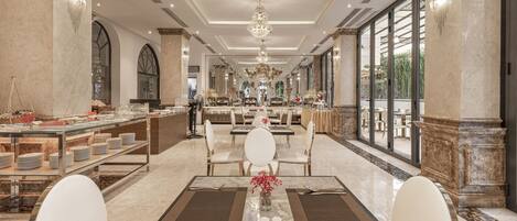 A spacious hotel lobby with a marble floor, a central dining area with white chairs, a buffet with various food items, and large windows offering a view of the outdoors.