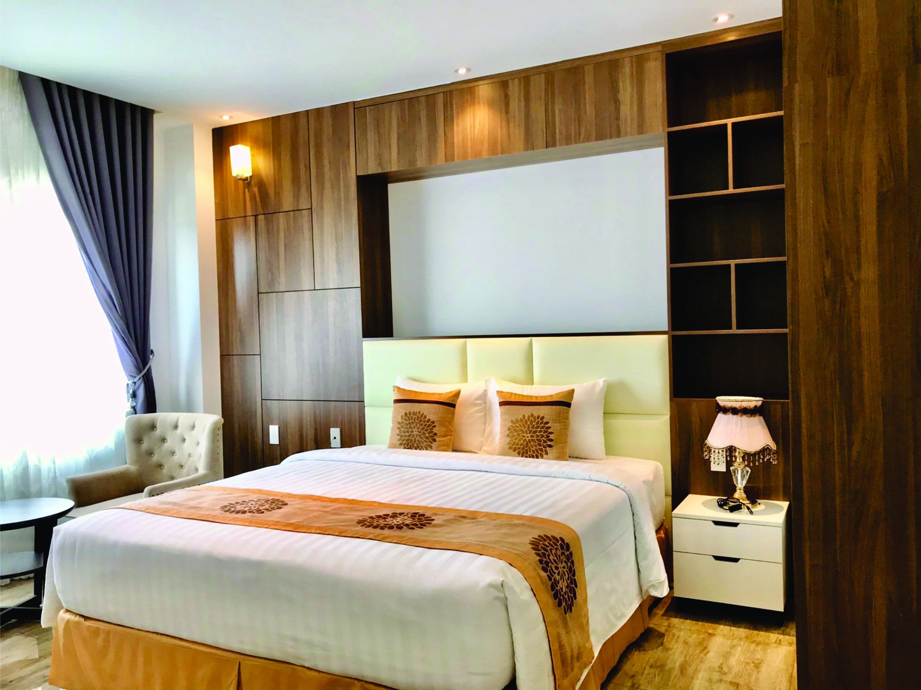 A hotel room with a large bed, a bedside table with a lamp, a chair, and a built-in shelving unit.