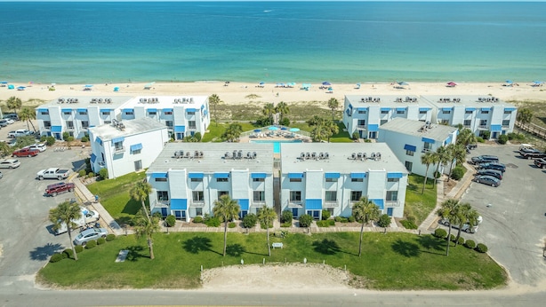 Un hôtel situé en bord de mer, composé de plusieurs bâtiments aux couleurs bleu et blanc, avec un parking et une plage de sable dotée de parasols.