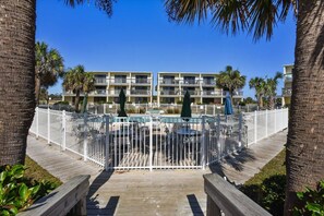 A resort with a pool area surrounded by a white fence, palm trees, and a multi-story building with balconies.