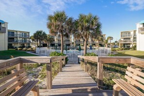 A wooden boardwalk leading to a pool area with palm trees and a clear sky.