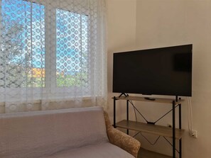 Una habitación con una televisión sobre un mueble bajo, una ventana con cortinas de encaje y un sofá.