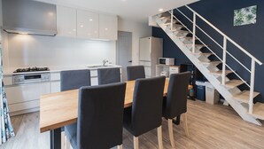 A modern kitchen with white cabinets, a wooden dining table, and black chairs. There is a staircase with white railings leading to a dark wall with a tropical print. - Villa Garden Onna (Onna)