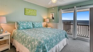 A bedroom with a bed, a ceiling fan, a nightstand, a lamp, a wall art piece, and a view of the ocean through a balcony.