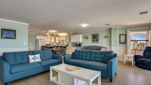 A living room with two blue sofas, a white coffee table, and a kitchen in the background.
