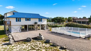 Une maison à deux étages avec un toit bleu, une terrasse couverte et une piscine entourée d’une clôture blanche.