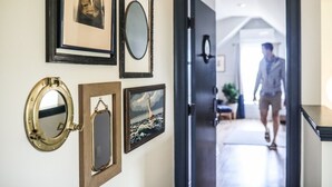 Un pasillo con una puerta que da a una habitación. En la pared hay varios objetos enmarcados, incluyendo un retrato, un espejo circular y una pintura de paisaje.
