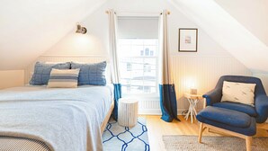 Un dormitorio con cama, una silla, una mesita y una ventana con cortinas.