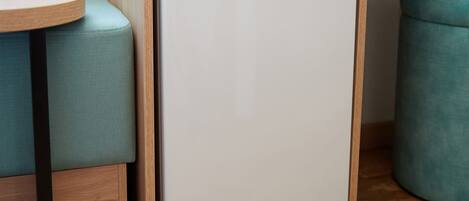 A compact white refrigerator in a corner of a room with teal upholstered seating and wooden flooring.