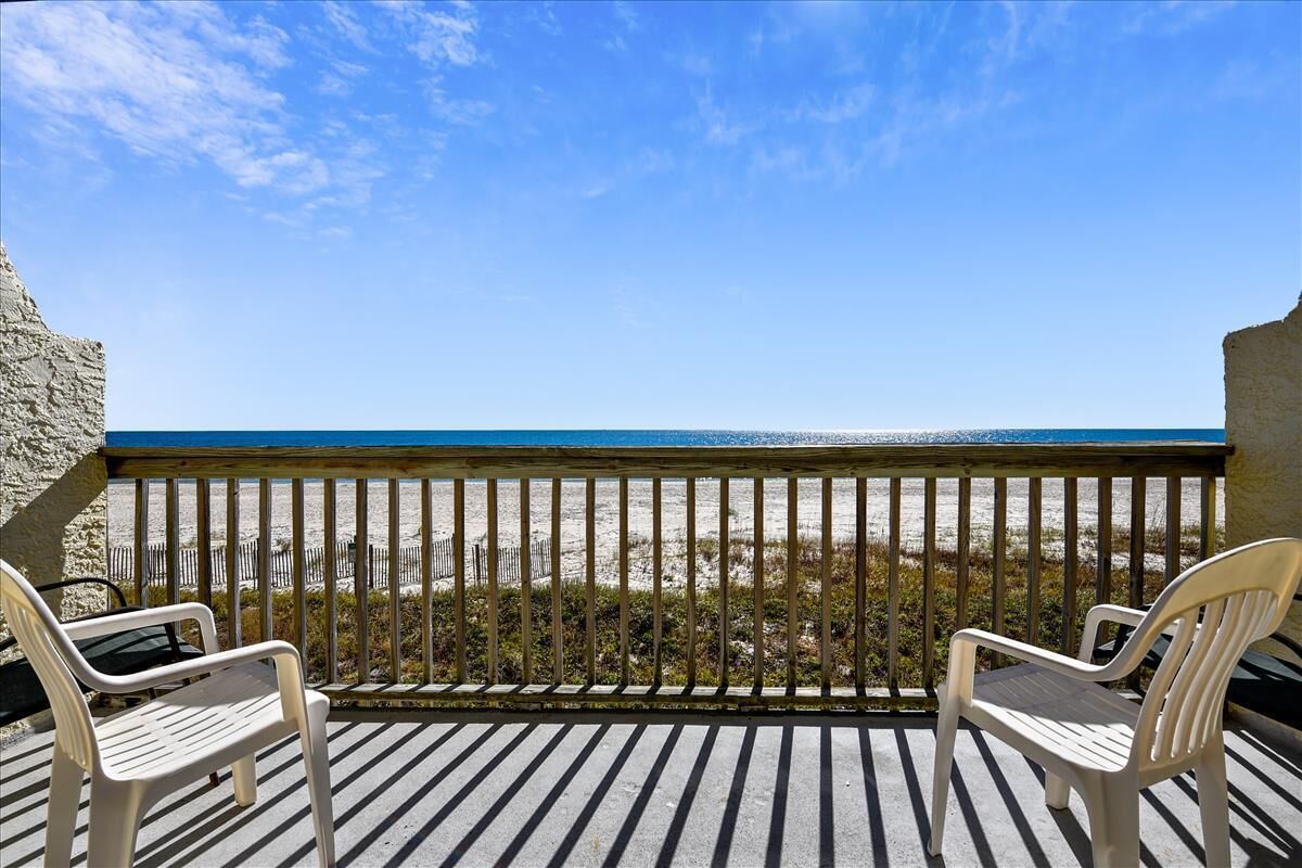 A balcony with two white plastic chairs, a wooden railing, and a view of the beach.