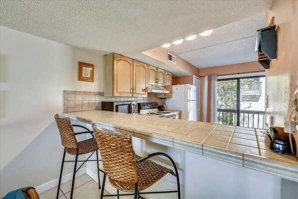 A kitchen with a long island counter, two wicker bar stools, wooden cabinets, and a window with a view.