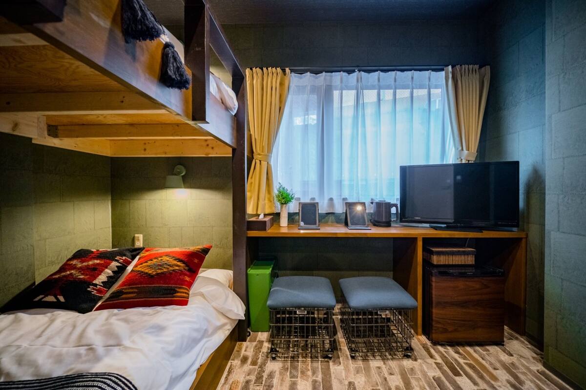 Habitación pequeña con literas, un escritorio con ordenador y televisión, y una ventana con cortinas.