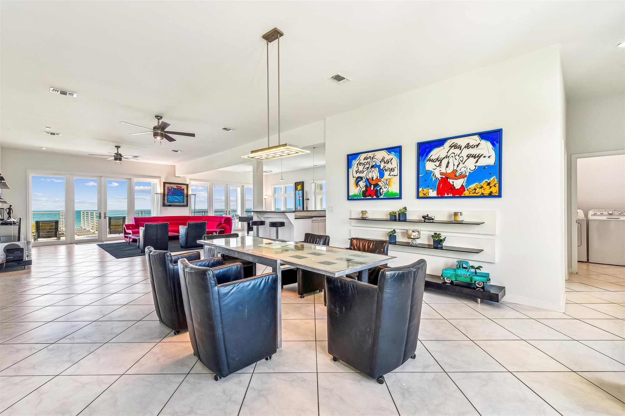 A spacious living area with a dining table, chairs, and a red sofa. There are ceiling fans, a kitchen area, and artwork on the walls.