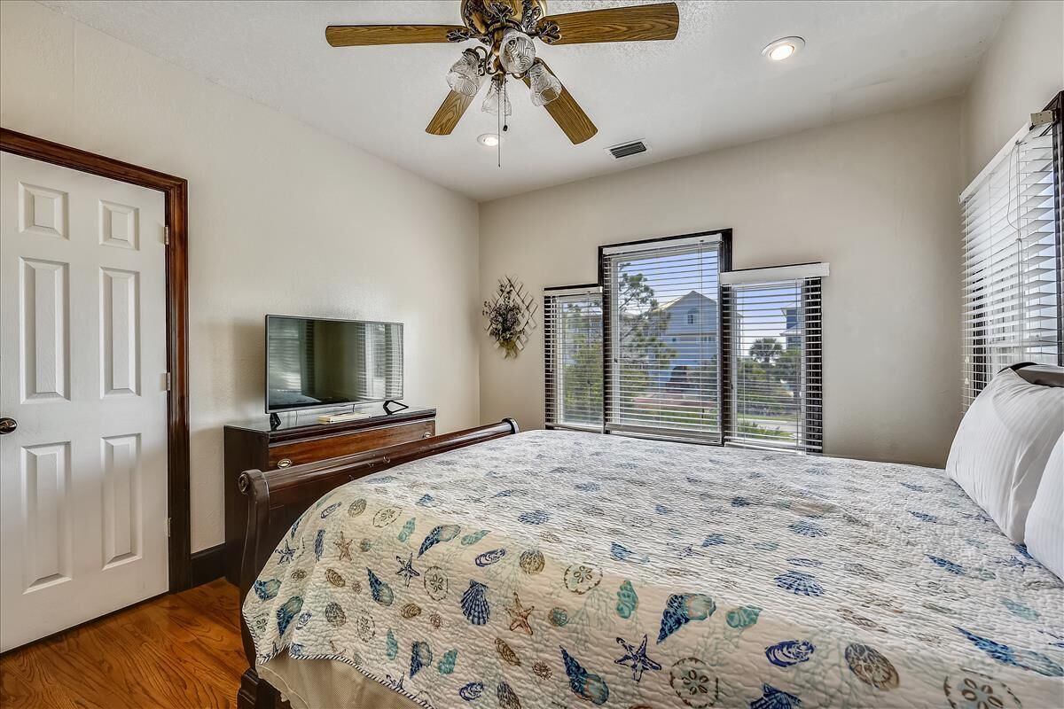 A bedroom with a bed, a ceiling fan, a television on a dresser, and a window with blinds.