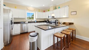 A modern kitchen with white cabinets, a stainless steel refrigerator, a large island with stools, and a window with a view.