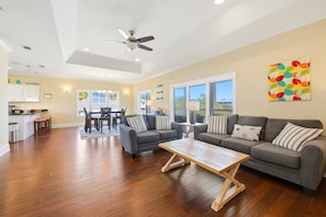Uma sala de estar ampla com piso de madeira, um conjunto de sofá cinza, uma mesa de centro de madeira e um ventilador de teto.