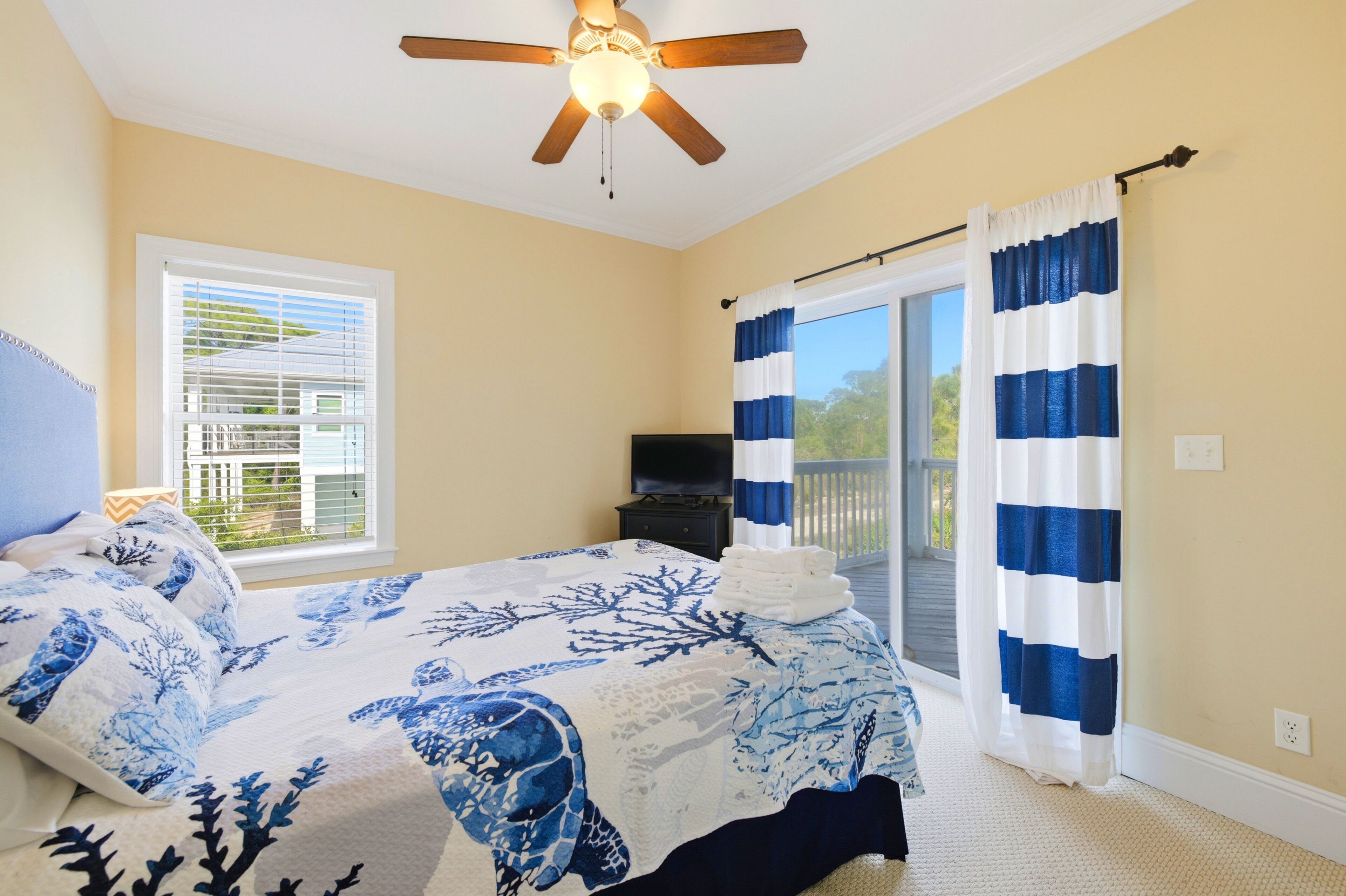 A bedroom with a bed, a ceiling fan, a television, and a view through a window.