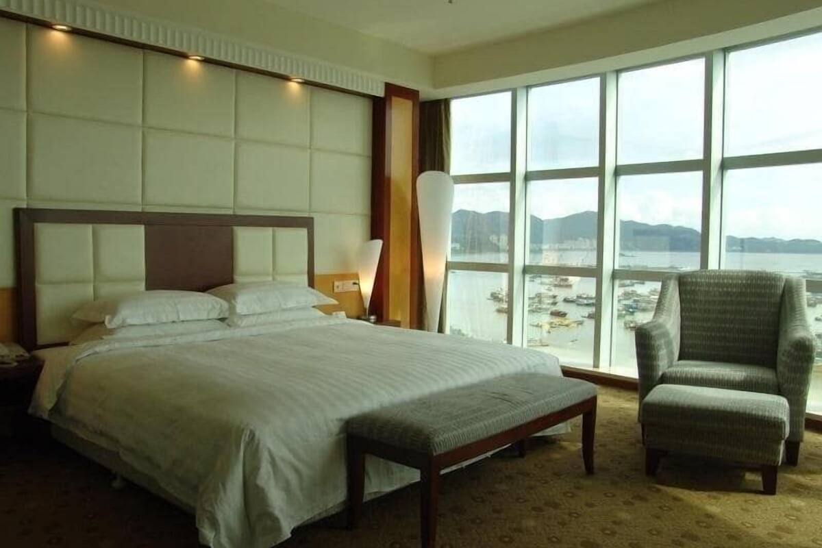 Een hotelkamer met een groot bed, een stoel en uitzicht op zee door grote ramen.
