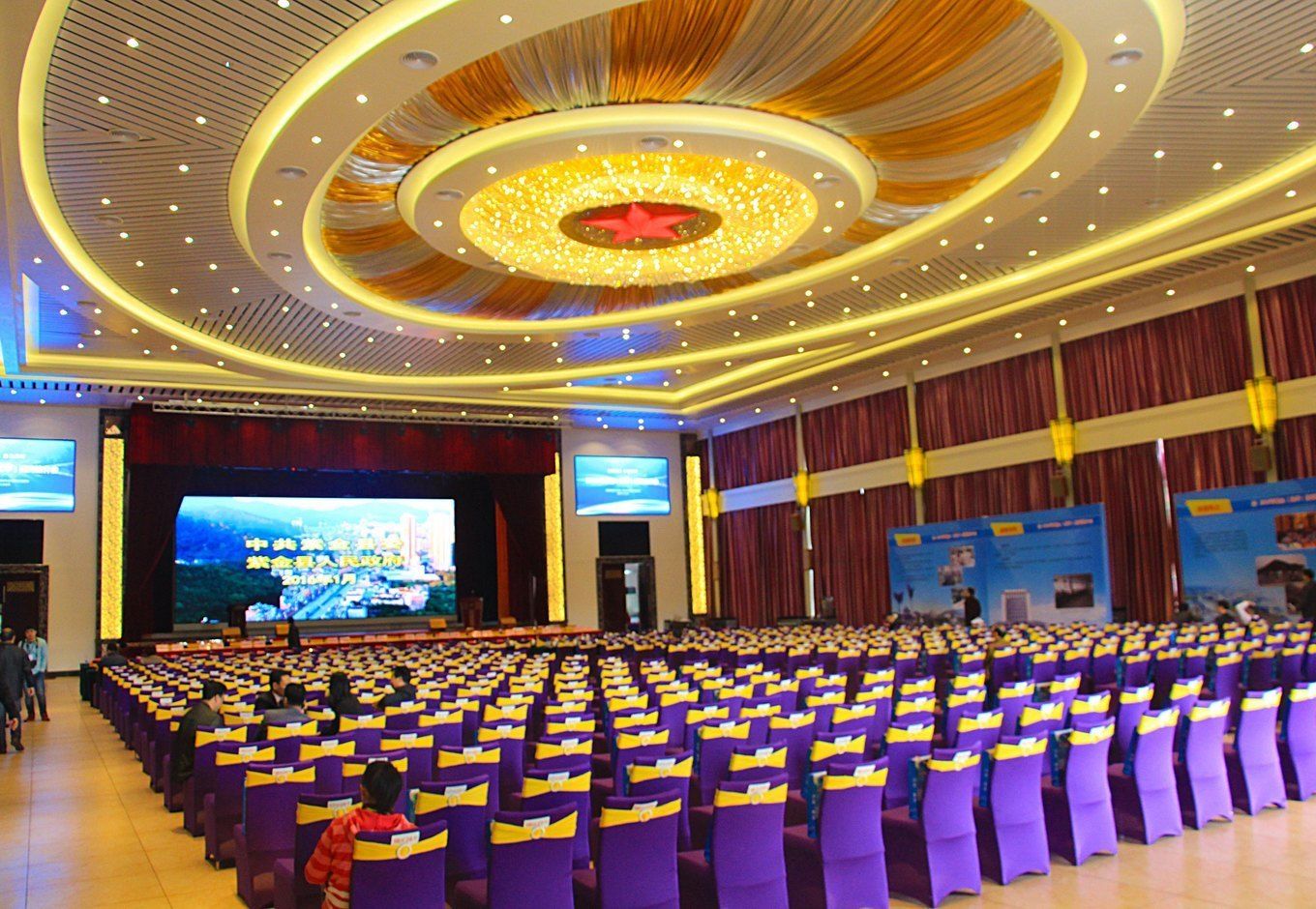 A large auditorium with rows of purple chairs, a central stage, and a circular ceiling design.
