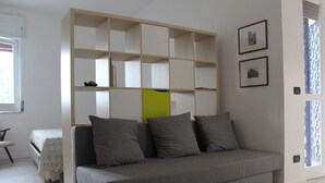 A modern living room with a grey sofa, a wooden bookshelf, and a bed with a floral bedspread. - PioppiHouse Casa Vacanza.  Large independent suite with small terrace overlooking the sea (Acciaroli)
