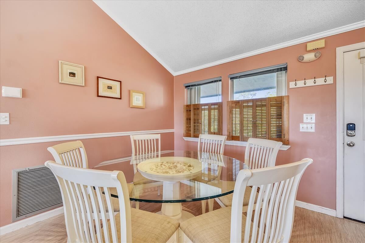 A dining area with a glass table surrounded by white chairs, framed pictures on the wall, and a window with blinds.