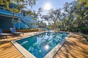 Una piscina amb terrassa de fusta i gandules, envoltada d'arbres i un edifici amb façana blava.