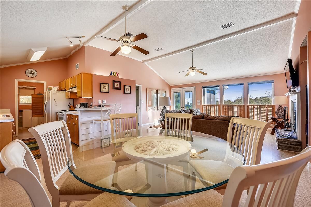 A spacious living area with a glass-top dining table surrounded by white chairs, a kitchen area with wooden cabinets, and a large window offering a view of the outdoors.