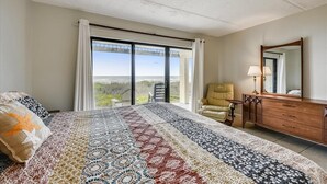 Habitación de hotel con una cama grande, un aparador de madera, una silla y vistas al exterior a través de una puerta corrediza.