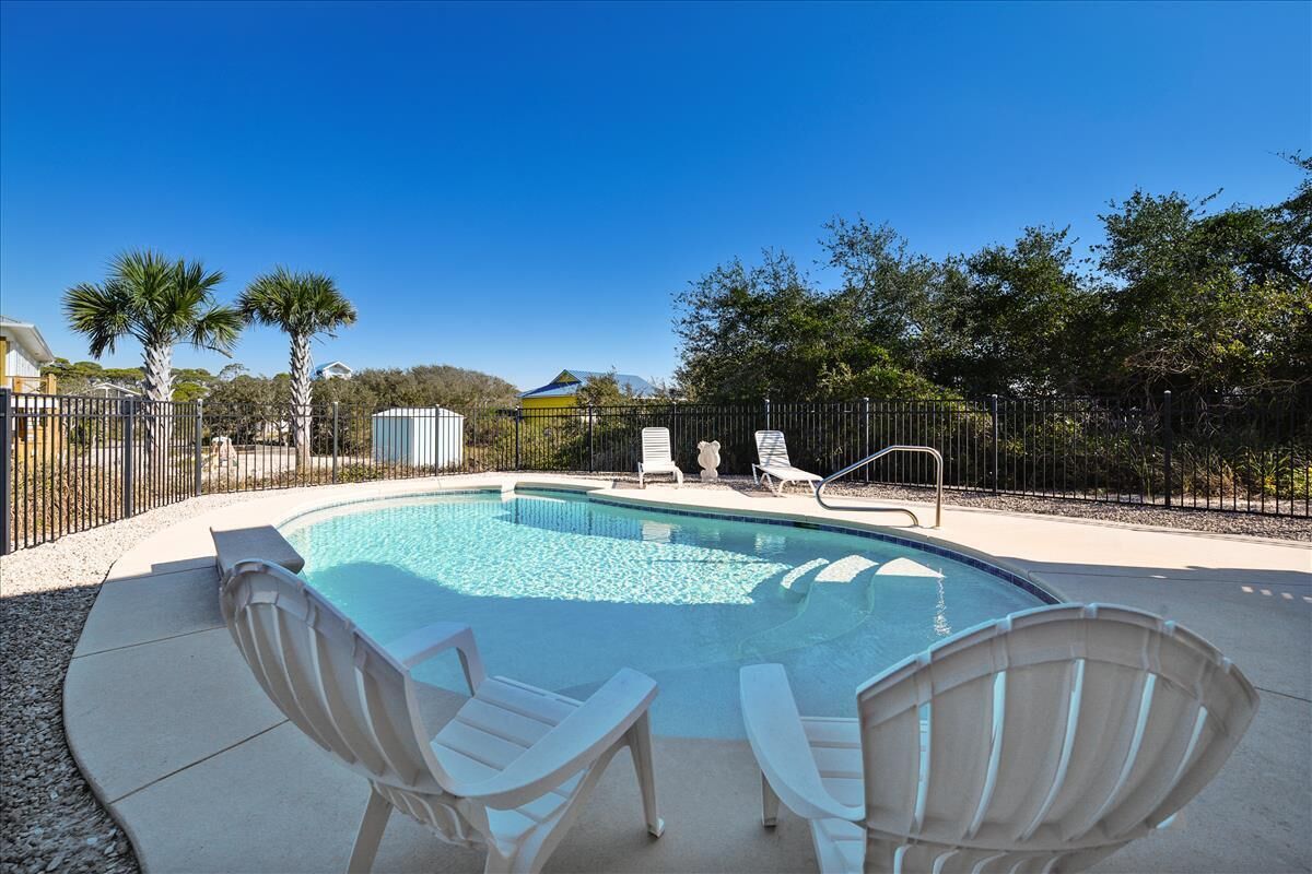 A swimming pool with four white lounge chairs, surrounded by a black fence and palm trees.