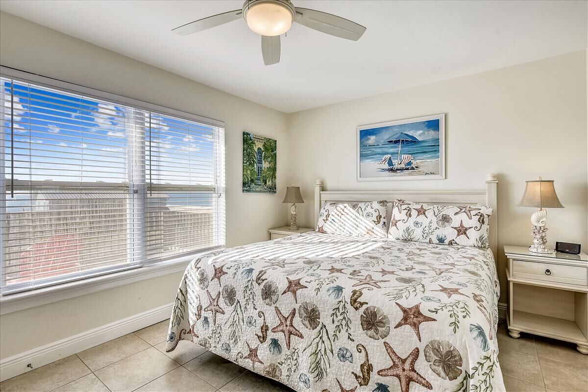 A bedroom with a bed, a ceiling fan, a window with blinds, a nightstand, and a painting on the wall.