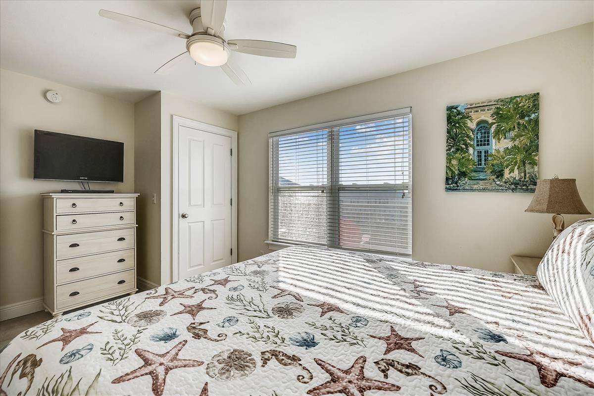 A bedroom with a bed, a dresser, a television, a ceiling fan, a window with blinds, and a painting on the wall.