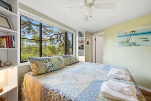 A bedroom with a large window, a ceiling fan, a bed with floral bedding, a bookshelf, and a painting of a beach scene. - BIRD WATCHER IS PARADISE FOR A NATURE LOVER!! (St. George Island)